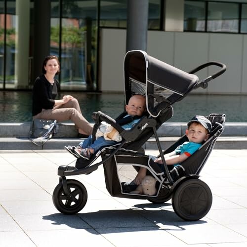 Deux enfants dans une poussette double avec une femme en arrière-plan.