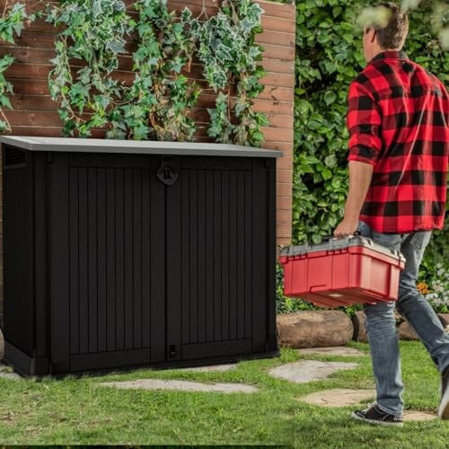Homme portant une boîte à outils devant un abri de jardin.